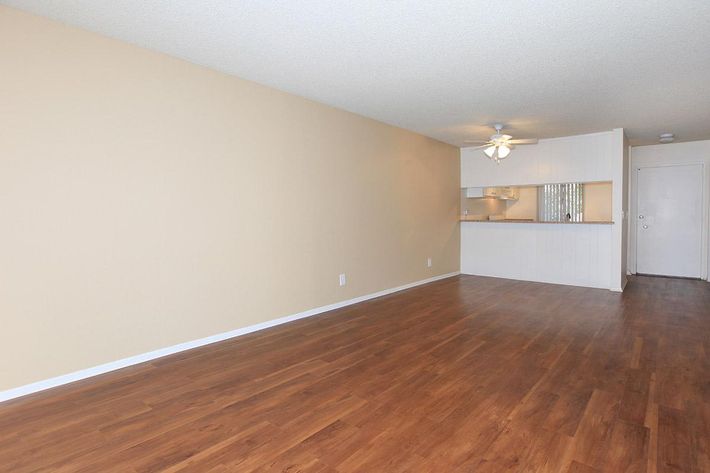 A spacious room with light beige walls and wooden flooring. There is a ceiling fan overhead, and an open kitchen area with white cabinetry in the background. The space is well-lit and features a doorway leading to the exterior, creating a bright and inviting atmosphere.