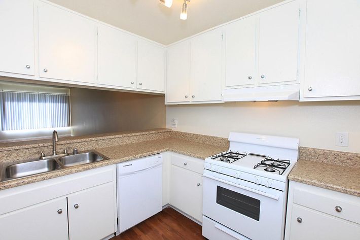 Modern kitchen featuring white cabinets, a granite countertop, a double sink, and a gas stove. Includes a built-in dishwasher and window with blinds providing natural light. The flooring is wooden, creating a warm ambiance. Room is well organized and designed for functionality.