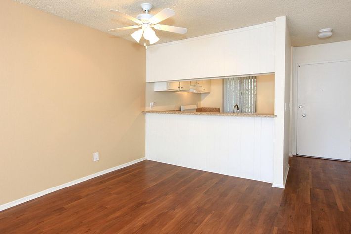 A bright, open living space featuring light beige walls and hardwood flooring. A ceiling fan is visible above, and there is a small bar area with a countertop separating it from a kitchen area in the background. The room has a simple design, with a door leading to the outside. Natural light shines through the space.