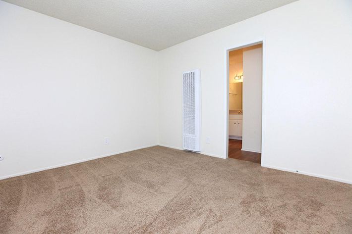 A vacant room with beige carpet flooring and light-colored walls. There's a white heating unit on the left side and an open doorway leading to a bathroom area in the background. Natural light fills the space, giving it an airy feel. The room is unfurnished and designed for versatile use.