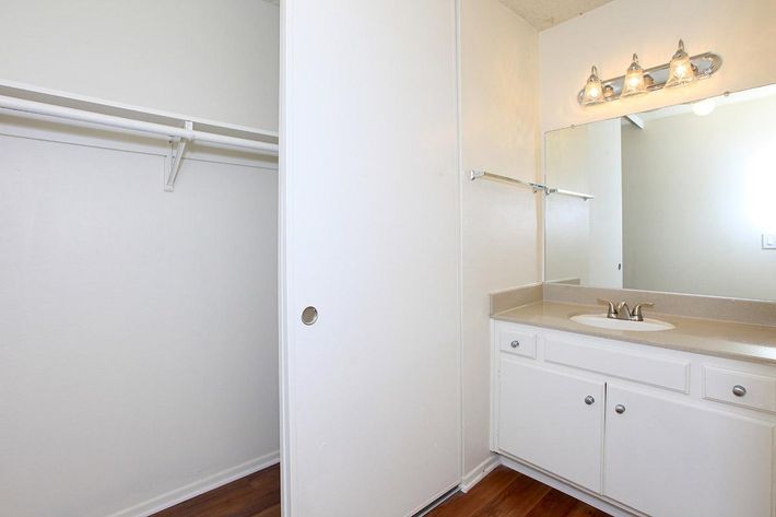 A clean, minimalistic bathroom featuring a small countertop with a sink, a mirror, and a light fixture above. There is a closet space to the left with open shelves, and the floor is made of hardwood. The walls are painted in a light color, creating a bright and airy atmosphere.