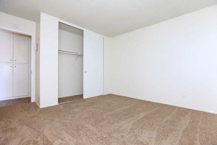 A spacious, empty room with beige carpet and plain white walls. There is a closet with sliding doors on one side, and a small wall with visible electrical outlets. The room has no furniture, giving it a clean and open appearance. Light possibly filters in from a window not shown in the image.