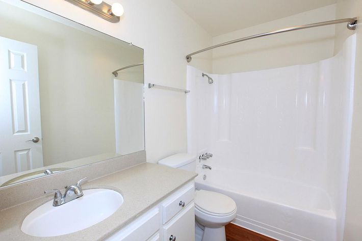 A clean, modern bathroom featuring a white bathtub with a curved shower rod, a toilet, and a double sink vanity with a countertop. A large mirror is mounted above the sink, and a door is visible in the background. The walls are painted a light color, creating a spacious feel.