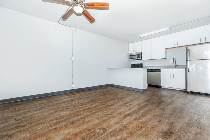 Interior view of an empty apartment showcasing an open layout. The space features wooden flooring, a ceiling fan, and a kitchen area equipped with white cabinets, a stainless steel refrigerator, and a microwave. The walls are light-colored, contributing to a bright and airy atmosphere.