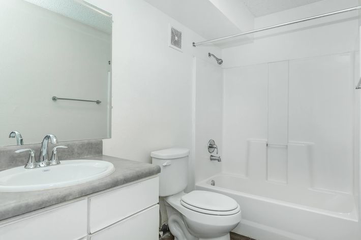 A clean, modern bathroom featuring a white bathtub and shower combination, a white toilet, and a sink with a silver faucet. The walls are light-colored, and there is a large mirror above the sink. A towel rack is mounted on the wall, and the floor is covered with dark tiles.