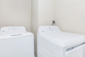 A white washing machine and a white dryer are positioned side by side in a laundry nook. The walls are a neutral color, and there is a small electrical outlet visible behind the appliances. The overall setting is clean and simple, emphasizing functionality.