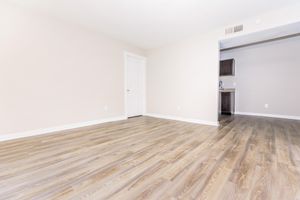 A spacious, empty room with light-colored walls and a hardwood floor. A closed white door is visible on the left side, and an entrance to a kitchen can be seen in the background. The room is well-lit, creating a clean and inviting atmosphere, perfect for decorating or furnishing.