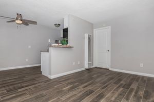 An interior view of a modern living space featuring smooth, dark wood flooring, a light gray wall, and a ceiling fan. To the left, a small kitchenette is visible with a bar area, while a door leads to another room. Natural light highlights the clean and minimalist design of the area.