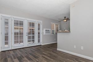 Bright and airy interior of a modern apartment featuring light gray walls, wood-style flooring, and large windows with white shutters. A ceiling fan hangs above the bar area, which is adjacent to an air conditioning unit. The space is well-lit, creating a welcoming atmosphere.