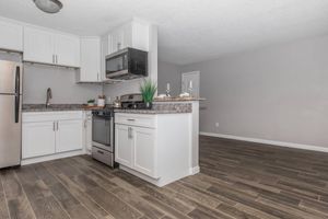A modern kitchen featuring white cabinets, a stainless steel refrigerator, and microwave. The kitchen island has a stone countertop with decorative items, including glasses and a small plant. The floor is covered with dark wooden tiles, and the walls are painted in a light gray color, creating a clean and inviting atmosphere.