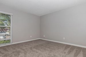 An empty room with light gray walls and a plush carpet. A window with white blinds lets in natural light, revealing a view of greenery outside. The space is clean and minimalistic, ideal for personal customization.