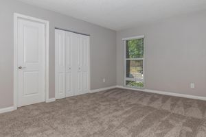 A spacious and well-lit room featuring light gray walls, plush carpet flooring, and a large window with blinds. It includes a closet with double doors and a plain white door leading to another area. The room has a cozy, inviting atmosphere with natural light streaming in.