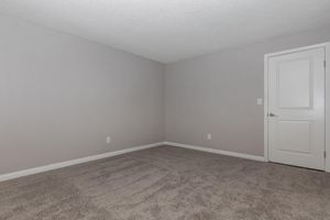 A plain, empty room with light gray walls and a white door. The carpet is light beige and appears soft. There are no furnishings or decorations, giving the space a minimalist look. The room is well-lit, with a textured ceiling.