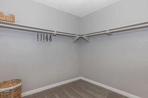 An empty closet with light gray walls and carpeted floor. It features two shelves installed on opposite walls, and there are a few black hangers hanging from one shelf. A woven basket and a laundry basket sit in the corner, indicating a neat and organized space.