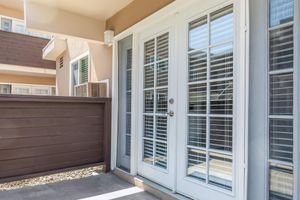 A close-up view of a set of white French doors with window panes, leading to an outdoor area. The doors are flanked by neutral-colored walls and feature a wooden railing. Soft sunlight casts shadows on the ground, creating a warm ambiance in a modern architectural setting.