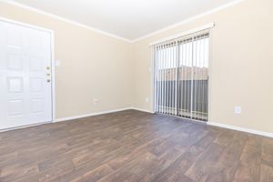 A spacious, empty room with light-colored walls and dark wood-like flooring. The entrance door is on the left, and a large window with vertical blinds is on the right, letting in natural light. The room has a clean, minimalist look, ideal for various decor styles.