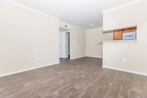 A spacious, unfurnished living area with beige walls and wood-like flooring. There's a kitchen counter with cabinets in the background, and a closed door leading to another room. The lighting is bright, creating an inviting atmosphere.