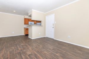 A vacant room featuring light-colored walls, a wood-like floor, and a kitchen area with wooden cabinets. The kitchen is partially visible, showing a microwave and limited appliances. A white door leads outside, and natural light illuminates the space.
