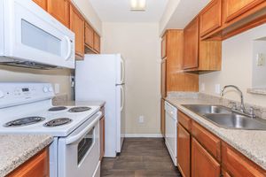 A modern kitchen featuring wooden cabinets, a double sink, and stainless steel appliances. The layout includes a white stove, microwave, and refrigerator. Countertops are patterned with light granite, and the space is well-lit, creating a warm and inviting atmosphere.