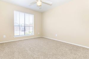 Empty room with beige walls and carpeted flooring, featuring a ceiling fan and a large window with blinds allowing natural light in. The room has minimal decor and no furniture, emphasizing a spacious and bright atmosphere.
