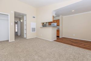A spacious living area featuring light beige walls and carpet, with an open layout connecting to a kitchen with wooden cabinets. There are doorways leading to other rooms, along with a small bar counter. Natural light brightens the space, creating a welcoming atmosphere.