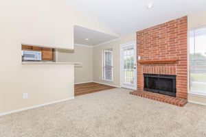 Living room with beige walls and carpet, featuring a brick fireplace. There's a small breakfast bar connecting to a kitchen area with a microwave visible. Large windows provide natural light, and there's a glass door leading outside, enhancing the airy feel of the space.