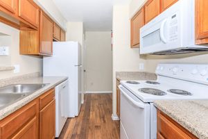 A bright and modern kitchen featuring wooden cabinets, a white refrigerator, a white microwave above a stove, and dual sinks. The countertops are a light granite, and the flooring is a warm hardwood. The layout is compact and functional, with a doorway visible leading to another area.