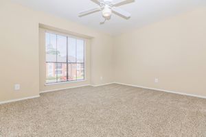 Empty room with beige walls and light brown carpet. A ceiling fan is mounted on the ceiling, and a window with blinds allows natural light to enter. The view outside shows a courtyard or other buildings. The space is simple and spacious, suitable for various furnishings or designs.