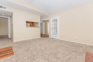 Spacious living area featuring light beige walls, plush carpet flooring, and a vaulted ceiling. An opening leads to a kitchenette with wood accents, and a door opens to an outdoor space. Natural light filters through the sliding glass door, creating an inviting atmosphere.