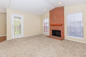 Cozy living room with a light beige color scheme, featuring a red brick fireplace, large windows with blinds, and plush carpet flooring. A glass door leads to an outdoor area, offering natural light and an inviting atmosphere.