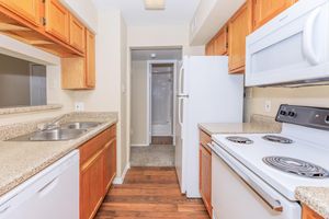 A view of a modern kitchen featuring wooden cabinets, granite countertops, a double sink, a dishwasher, a white refrigerator, and an electric stove. The hallway leads to a bathroom with a shower in the background, complemented by hardwood flooring throughout.