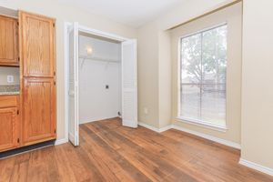 A bright and spacious room featuring hardwood floors, a large window with blinds, and a well-organized closet with sliding doors. The walls are painted in a light color, and there are wooden cabinets visible in the adjacent area, adding to the inviting atmosphere.