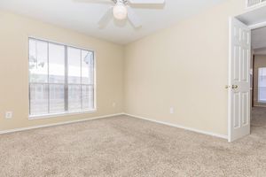 A well-lit, empty room featuring beige walls and carpet, with a window allowing natural light and a ceiling fan. A partially open white door leads to another area. The space has a clean and minimalist feel, perfect for personalizing or furnishing.