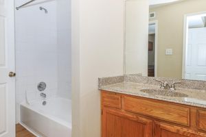 A clean bathroom featuring a bathtub with a showerhead, a light-colored wall, and a wooden vanity with a granite countertop. The mirror reflects the bathroom space, and a white door is visible in the background, adding to the overall spacious feel of the area.