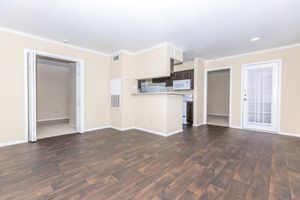 A spacious, well-lit apartment interior featuring hardwood-style flooring, beige walls, and a kitchenette with white appliances. The layout includes an open living area, connecting to a small room, and a sliding glass door leading to an outdoor space.