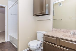 A clean, modern bathroom featuring a white toilet, a granite countertop with a sink, and dark brown cabinetry. A rectangular mirror hangs above the sink, and there is a bathtub with a shower rod in the background. The walls are a light beige color, and the floor has dark wood-like tiles.