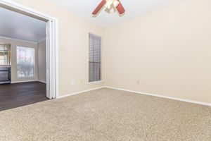 Empty room with light beige walls and carpet. A ceiling fan is visible, and there is a window with blinds. A doorway leads to another room in the background. The overall atmosphere is clean and neutral, suitable for various uses or personal decor.