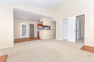 A spacious, light-filled apartment interior with beige walls and carpet. To the left, a living area features a large window and a brick fireplace. A doorway leads to the kitchen, which has wood cabinets and modern appliances. On the right, two doors open to a closet and another room. The layout emphasizes an open, inviting atmosphere.
