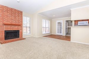 A spacious, well-lit living area featuring a brick fireplace, large windows, and light-colored walls. The room has plush carpeting and wood flooring leading to a kitchen area with modern appliances. A front door with glass panels adds to the inviting atmosphere.