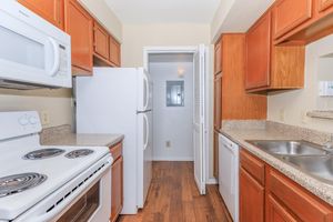 A view of a modern kitchen featuring wooden cabinetry, a white refrigerator, a white microwave, and a white stove with an oven. The countertops are granite, and the space has a hardwood floor. A doorway leads to another room, and there's a sink with a dishwasher nearby.