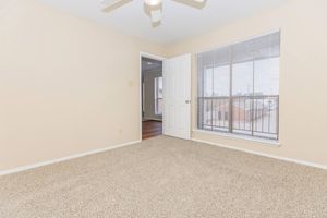 A light-colored empty room with beige walls and a tan carpet. A ceiling fan is visible, and there is a large window with white blinds. The door to another room is partially open, revealing a glimpse of hardwood flooring in the adjoining space. Natural light flows into the room, enhancing its airy feel.