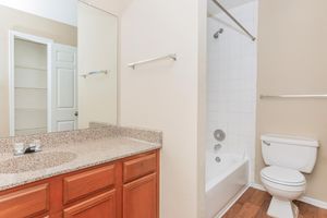 A clean and well-lit bathroom featuring a bathtub with a shower, a white toilet, and a wooden vanity with a granite countertop. The walls are painted in neutral tones, and there's a mirror above the sink. A closet door is visible in the background. The floor is covered in light wood laminate.