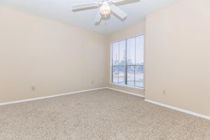 A small, empty room with beige walls and light brown carpet. There is a large window with blinds, allowing natural light to enter. The ceiling features a white ceiling fan. The space is unadorned and ready for furniture placement, creating a simple and open atmosphere.