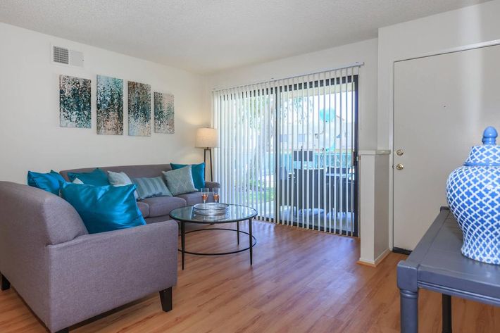 A cozy living room featuring a gray sofa adorned with blue and teal throw pillows. There's a round glass coffee table in front, and large sliding doors with vertical blinds open to a patio area. The walls are decorated with abstract artwork, and a floor lamp provides soft lighting in the corner.