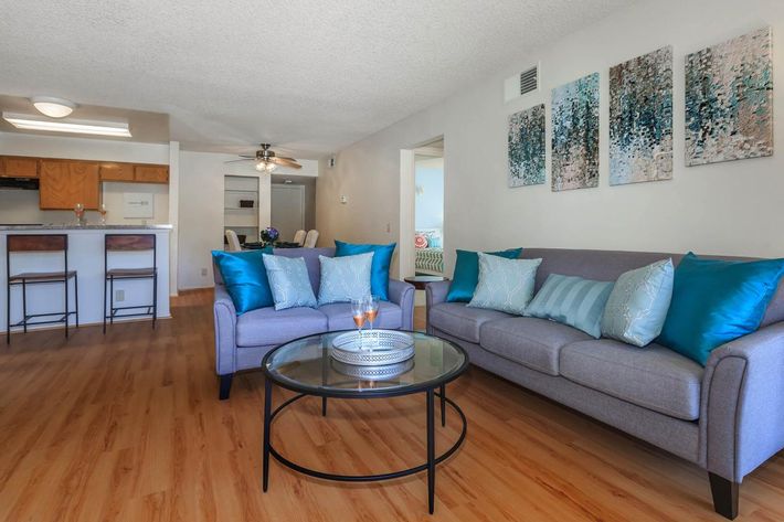 A cozy living room featuring a grey sofa with turquoise accent pillows, a glass coffee table, and a light wood floor. The space includes abstract wall art in shades of blue, a bar area with stools in the background, and an open layout connecting to a kitchen area. Bright and inviting ambiance.