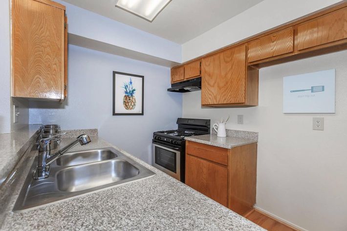 A modern kitchen featuring wooden cabinetry, a stainless steel sink with double bowls, a gas stove, and an oven. The countertops are made of granite, and there is a framed pineapple artwork on the wall. A wall fixture displays a cooking-related graphic. The floor is hardwood.