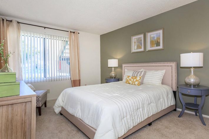 A cozy bedroom featuring a queen-sized bed with a light comforter, two decorative pillows, and a beige headboard. There are two bedside tables with lamps, a small seating area by the window with a chair, and light-colored curtains. The walls are painted a soothing green, and framed artwork hangs above the bed.