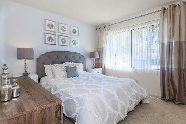 A cozy bedroom featuring a neatly made white quilted bed with decorative pillows, two bedside lamps on matching tables, and a wooden dresser. Light floods in through large windows with vertical blinds, complemented by elegant curtains. Four framed decorative pieces adorn the wall above the bed, enhancing the tranquil atmosphere.