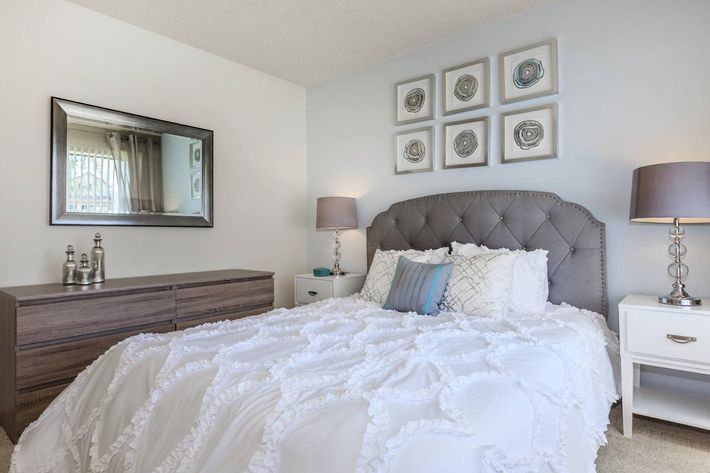A cozy bedroom featuring a large, tufted gray headboard with white bedding adorned with ruffled details. Two bedside tables with stylish lamps flank the bed. A modern mirror hangs on the wall, reflecting natural light, and four decorative framed artworks are arranged above the headboard. A wooden dresser completes the room.