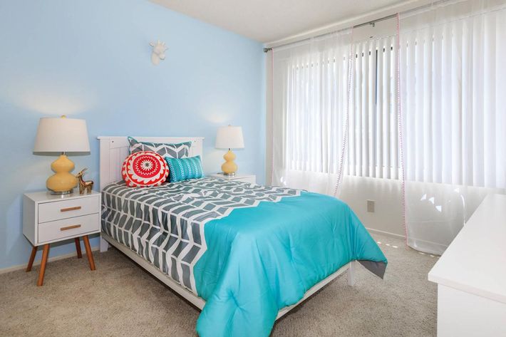A bright bedroom featuring a twin bed with a turquoise and gray patterned comforter. There are two lamps with yellow bases on either side of the bed, a colorful decorative pillow, and a large window covered with sheer white curtains. The room has light blue walls and a neutral carpet.